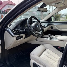 Auto Spa - Jasne wnętrze BMW X6 z czarnymi dywanikami, widok z zewnątrz przez otwarte drzwi kierowcy, dom w tle.