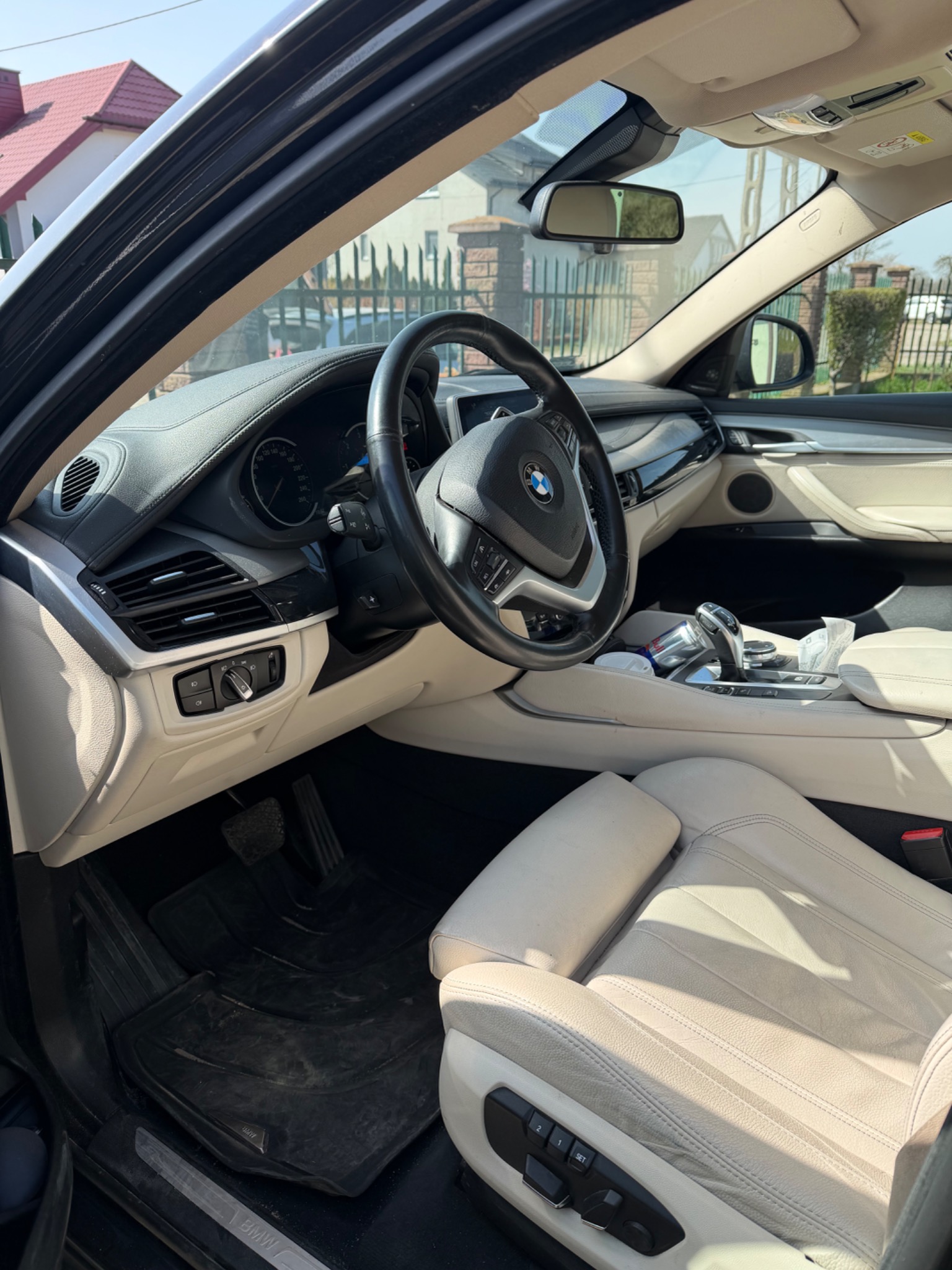 Jasne skórzane wnętrze BMW X6 z widocznymi detalami deski rozdzielczej, kierownicy i foteli. Widok z zewnątrz auta, w tle ogrodzenie i dom.