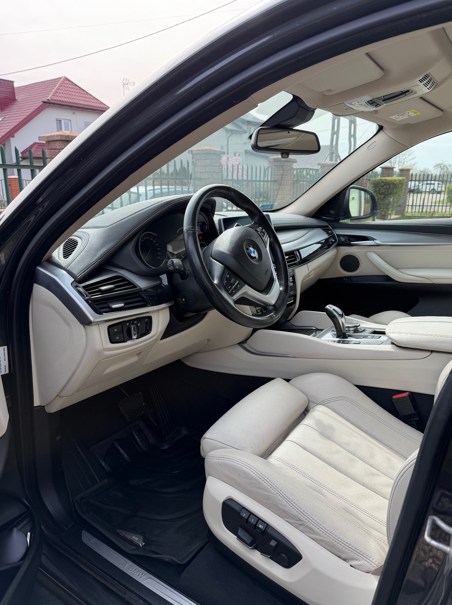 Jasne wnętrze BMW X6 z czarnymi dywanikami, widok z zewnątrz przez otwarte drzwi kierowcy, dom w tle.