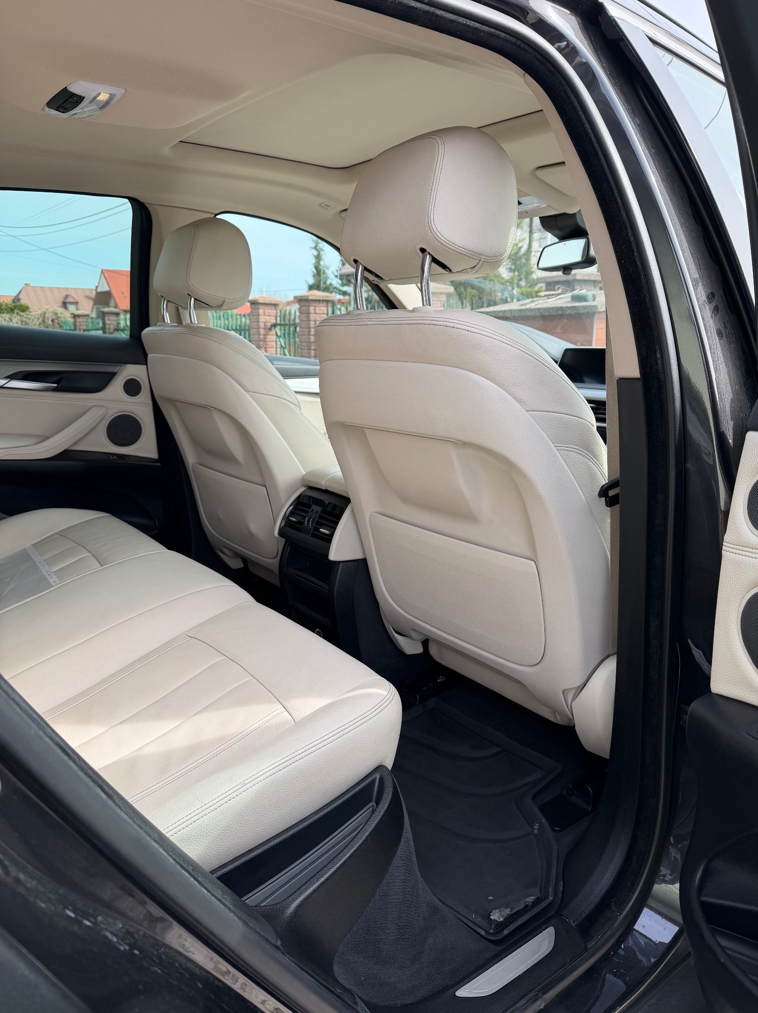 Jasne, skórzane fotele tylne w czarnym samochodzie SUV, widoczne przez otwarte drzwi. Czyste wnętrze, dywaniki gumowe. Widok na zewnątrz przez okno.