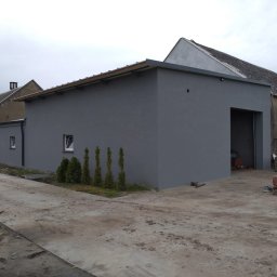 JAWI Projekty Budowlane Hubert Bryl - Szary budynek gospodarczy z płaskim dachem i otwartą bramą wjazdową, otoczony zielenią i betonowym placem. Widoczne są sąsiednie budynki i pochmurne niebo.