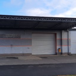 JAWI Projekty Budowlane Hubert Bryl - Szary budynek z bramą garażową i zadaszeniem wspartym na słupach. Na budynku widoczne okna z luksferów i reklama salonu meblowego.