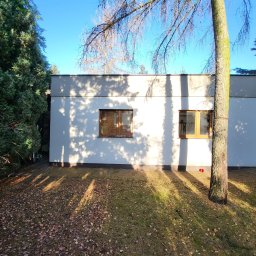 JAWI Projekty Budowlane Hubert Bryl - Nowoczesny, parterowy dom z płaskim dachem, oświetlony promieniami słońca, z widocznymi oknami i drzewem na pierwszym planie. Minimalistyczna architektura w otoczeniu zieleni.