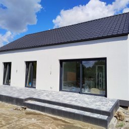 JAWI Projekty Budowlane Hubert Bryl - Nowoczesny, parterowy dom z szarą dachówką i wybrukowanym tarasem w Zaniemyślu. Widok z zewnątrz, w tle błękitne niebo z chmurami. Minimalistyczna architektura.