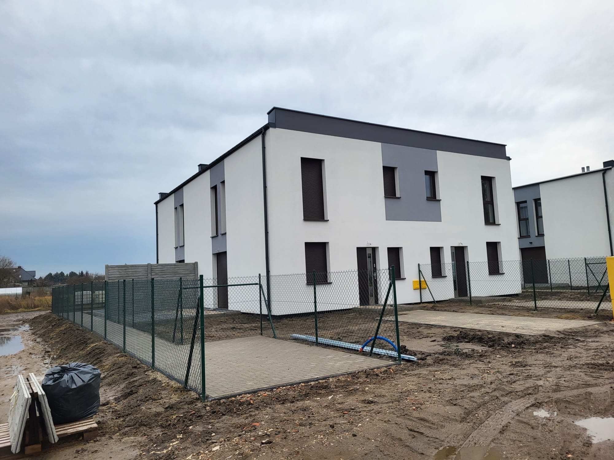 Nowoczesny, dwukondygnacyjny dom szeregowy z szarą elewacją i ciemnymi oknami, otoczony zielonym ogrodzeniem z siatki na błotnistym terenie. Pochmurne niebo w tle.
