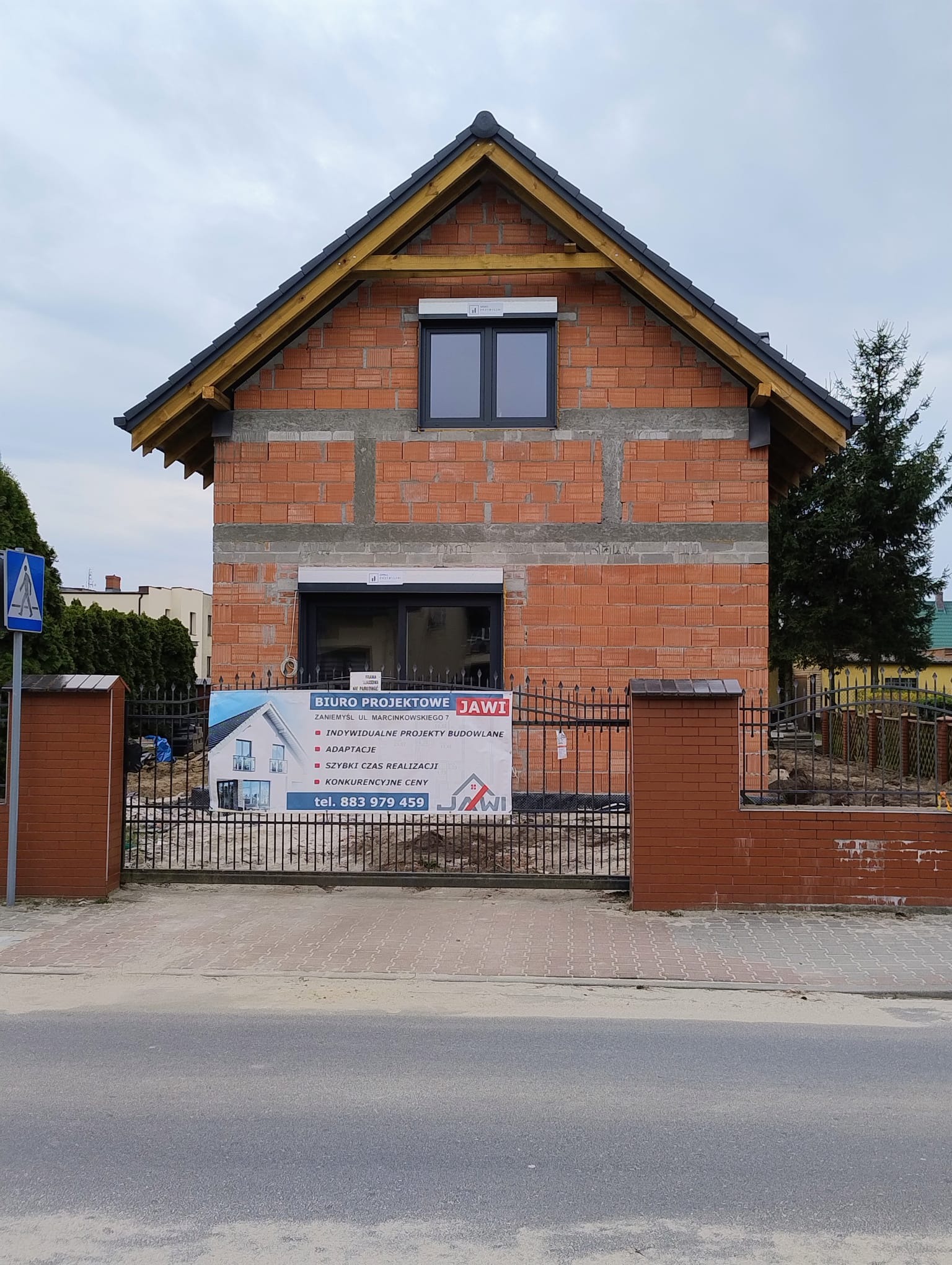 Surowy stan domu z czerwonej cegły, z widocznym banerem biura projektowego na ogrodzeniu. Okna zamontowane, dach w trakcie budowy. Widok z ulicy, pochmurne niebo.