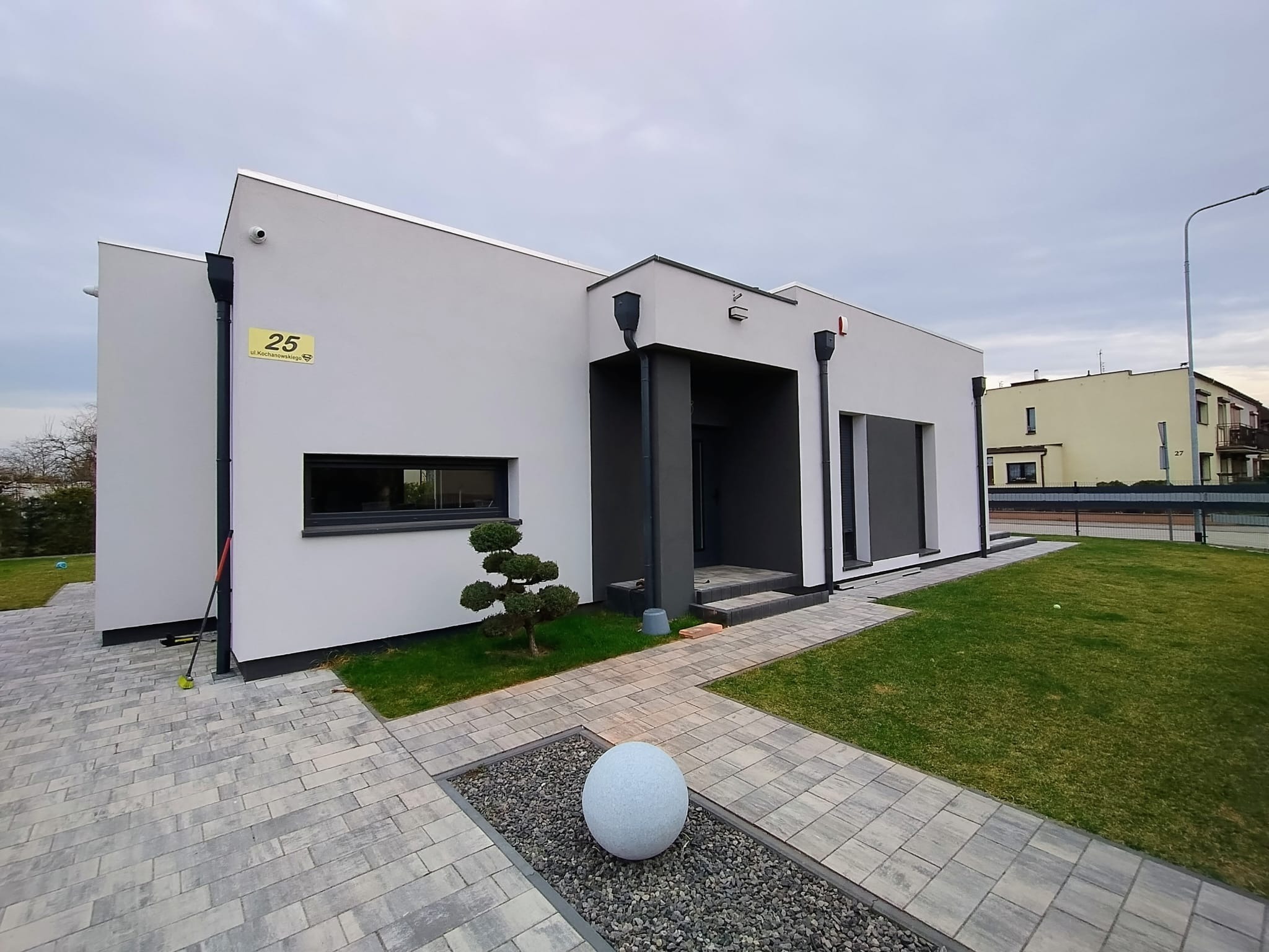 Nowoczesny, parterowy dom z płaskim dachem, numerem 25 na elewacji i ozdobnym drzewkiem bonsai w ogrodzie. Szara kostka brukowa prowadzi do wejścia.