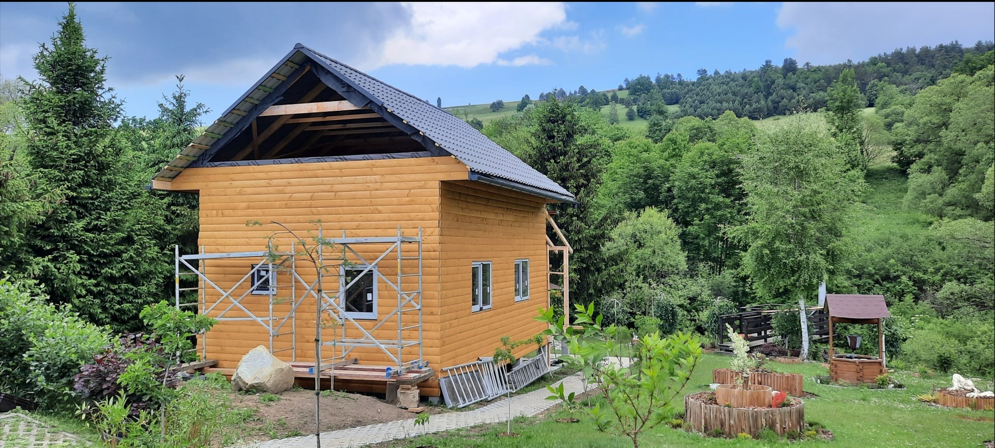 Drewniany dom w trakcie budowy z szarym dachem, otoczony zielenią. Widoczne rusztowania, drabiny i elementy ogrodu. W tle zalesione wzgórza pod błękitnym niebem.