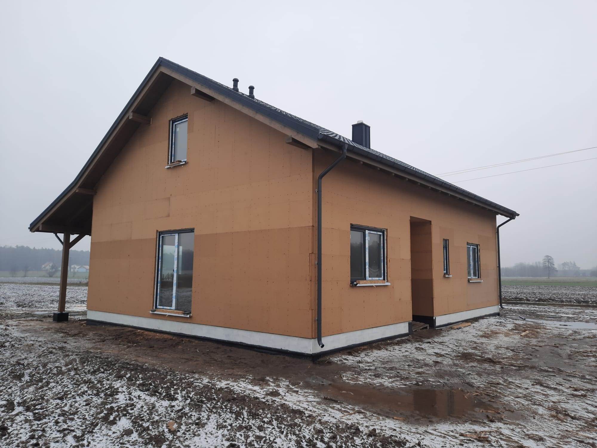 Surowy stan domu jednorodzinnego z drewnianą elewacją i czarnym dachem, widoczny w pochmurny dzień na tle zaśnieżonego pola. Okna wstawione, brak wykończenia.