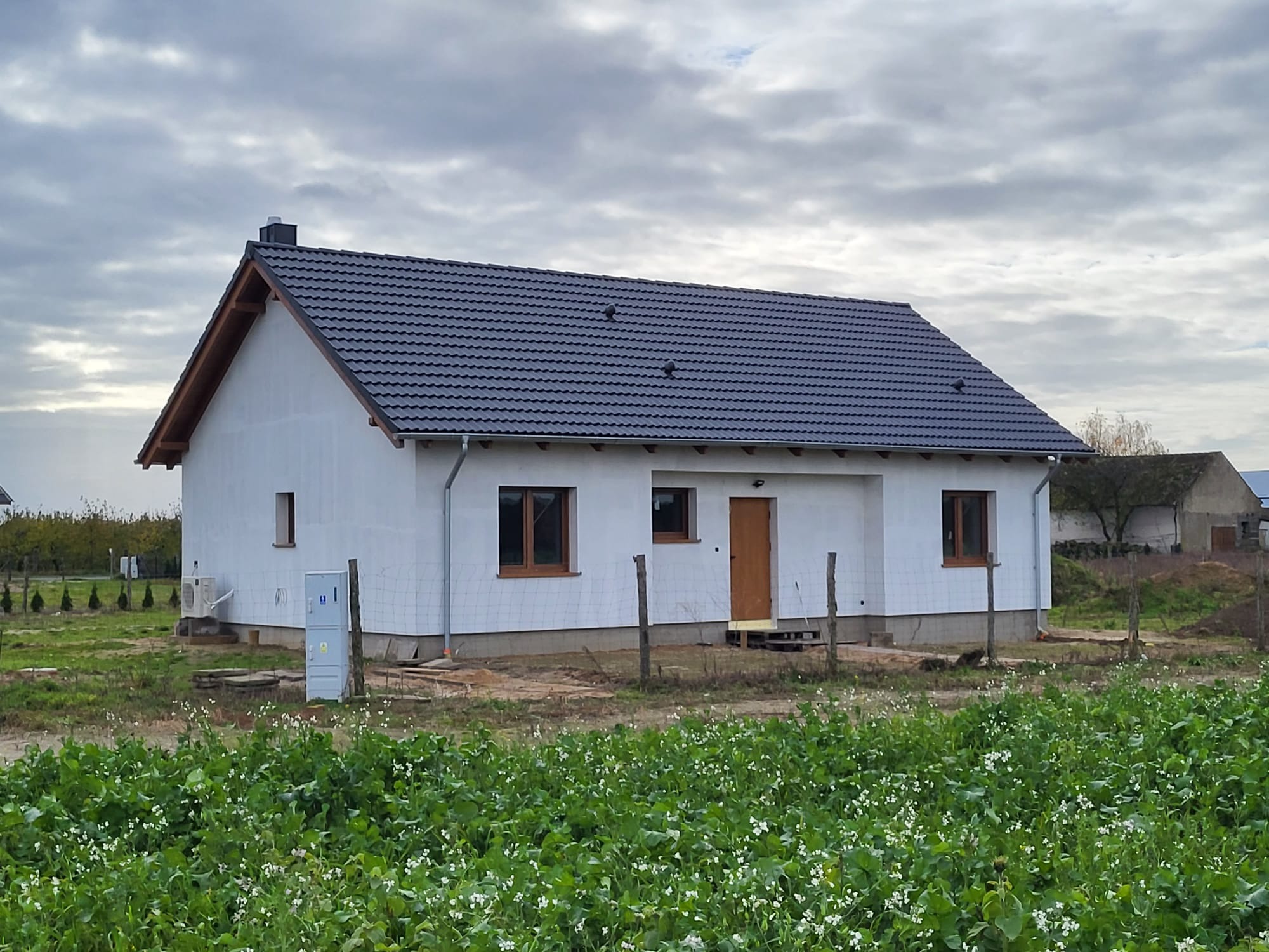 Niedawno wybudowany, parterowy dom z ciemną dachówką, jasną elewacją i brązowymi ramami okiennymi, otoczony zielenią i polnymi kwiatami w pochmurny dzień.