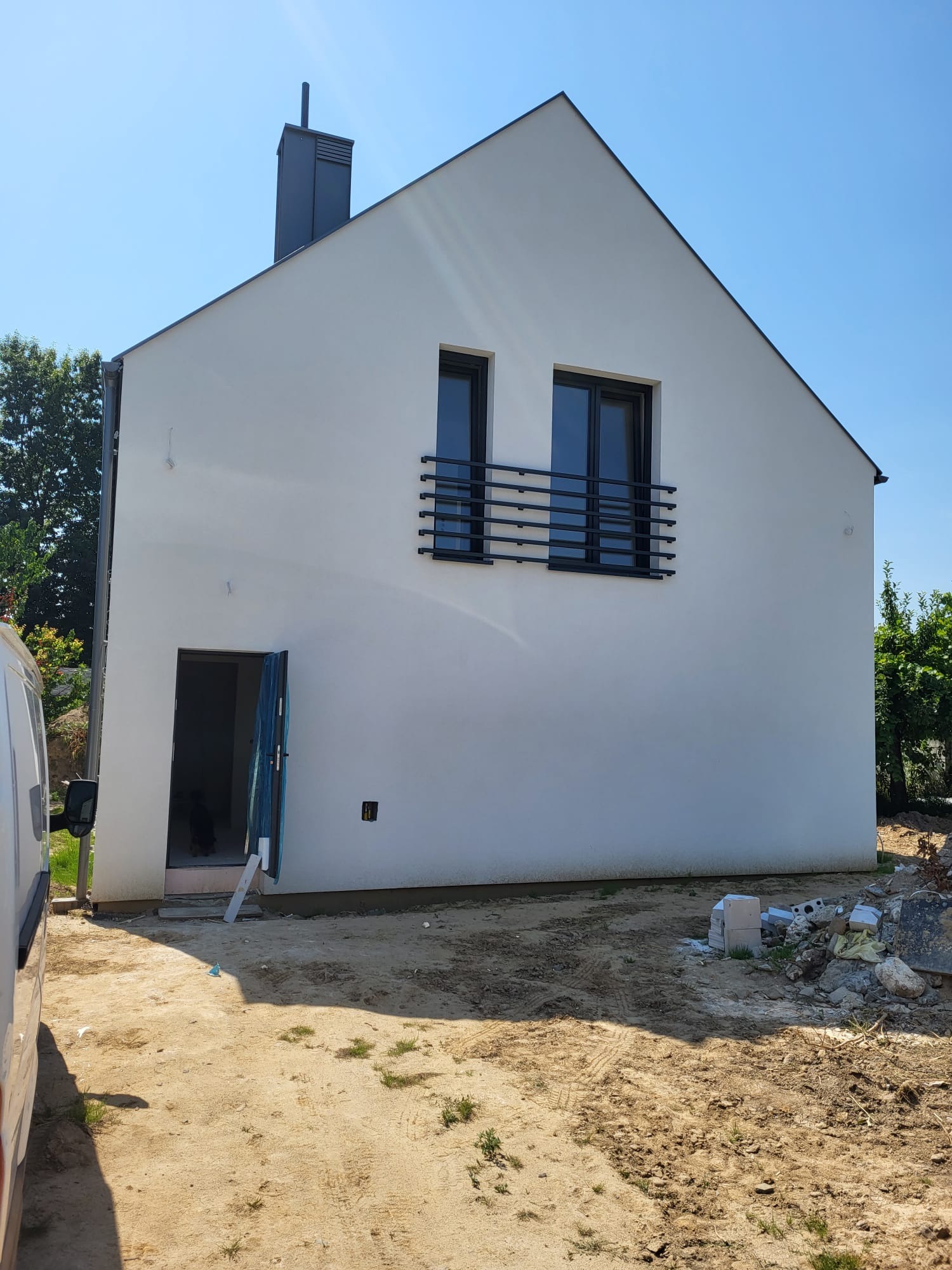 Biały dom jednorodzinny z czarnymi oknami i balkonem, widok z zewnątrz, teren wokół domu w trakcie prac wykończeniowych, błękitne niebo w tle.