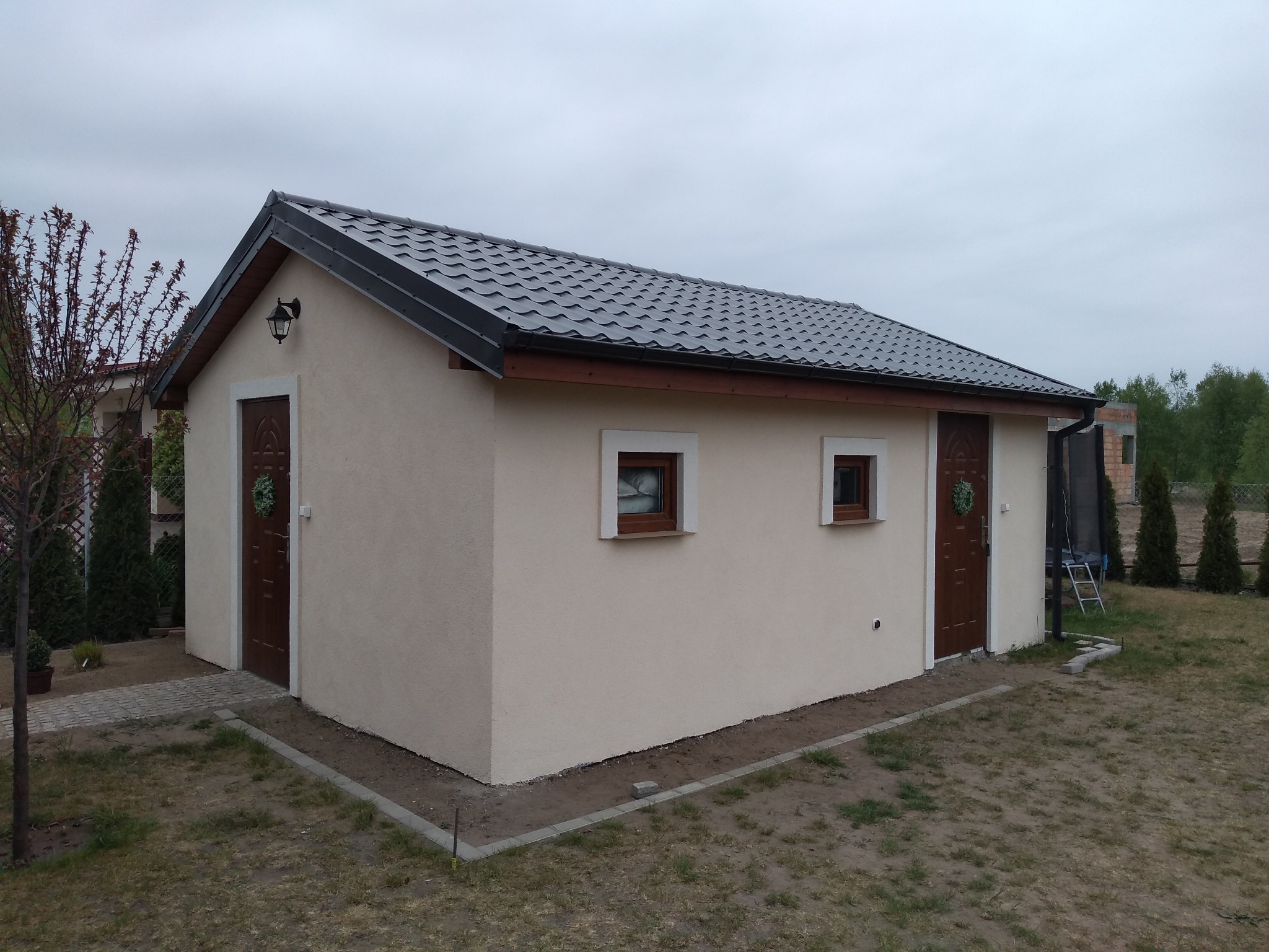 Gotowy, otynkowany budynek gospodarczy z dwuspadowym dachem z blachodachówki, dwoma wejściami z dekoracyjnymi wieńcami i dwoma oknami z drewnianą stolarką.