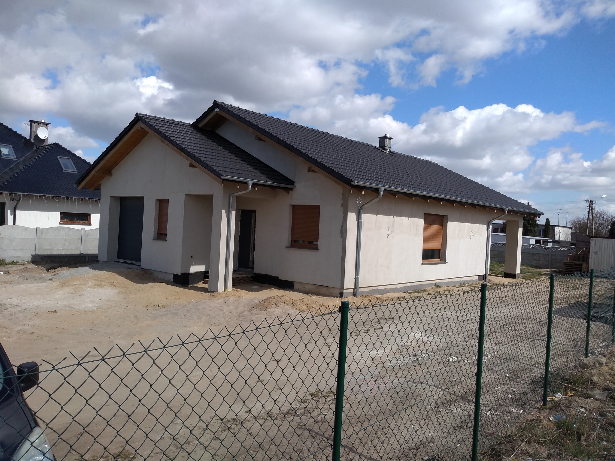 Niedokończony, parterowy dom z ciemną dachówką, widoczny zza zielonego ogrodzenia. Okna z roletami zewnętrznymi. W tle błękitne niebo z chmurami.