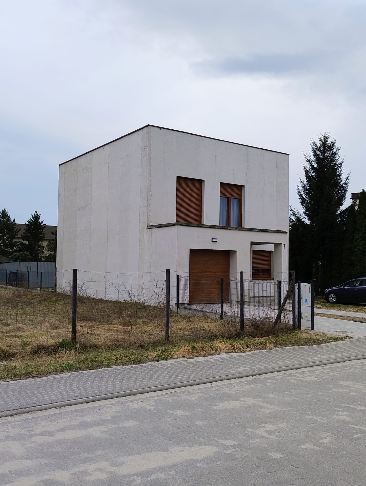 Nowoczesny, dwukondygnacyjny dom w kształcie sześcianu z brązowymi roletami, otoczony metalowym ogrodzeniem i suchą trawą. Minimalistyczna architektura.
