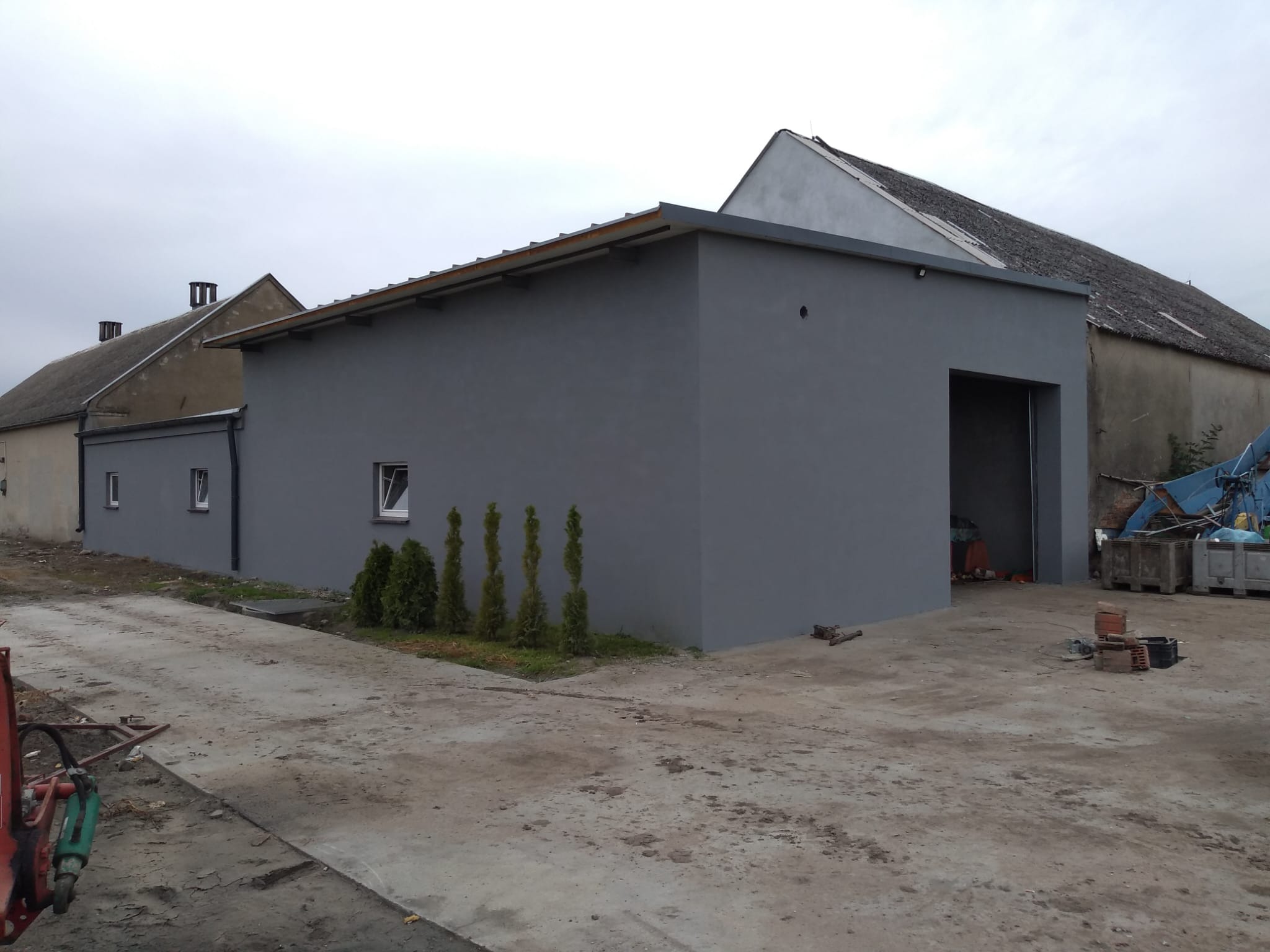 Szary budynek gospodarczy z płaskim dachem i otwartą bramą wjazdową, otoczony zielenią i betonowym placem. Widoczne są sąsiednie budynki i pochmurne niebo.