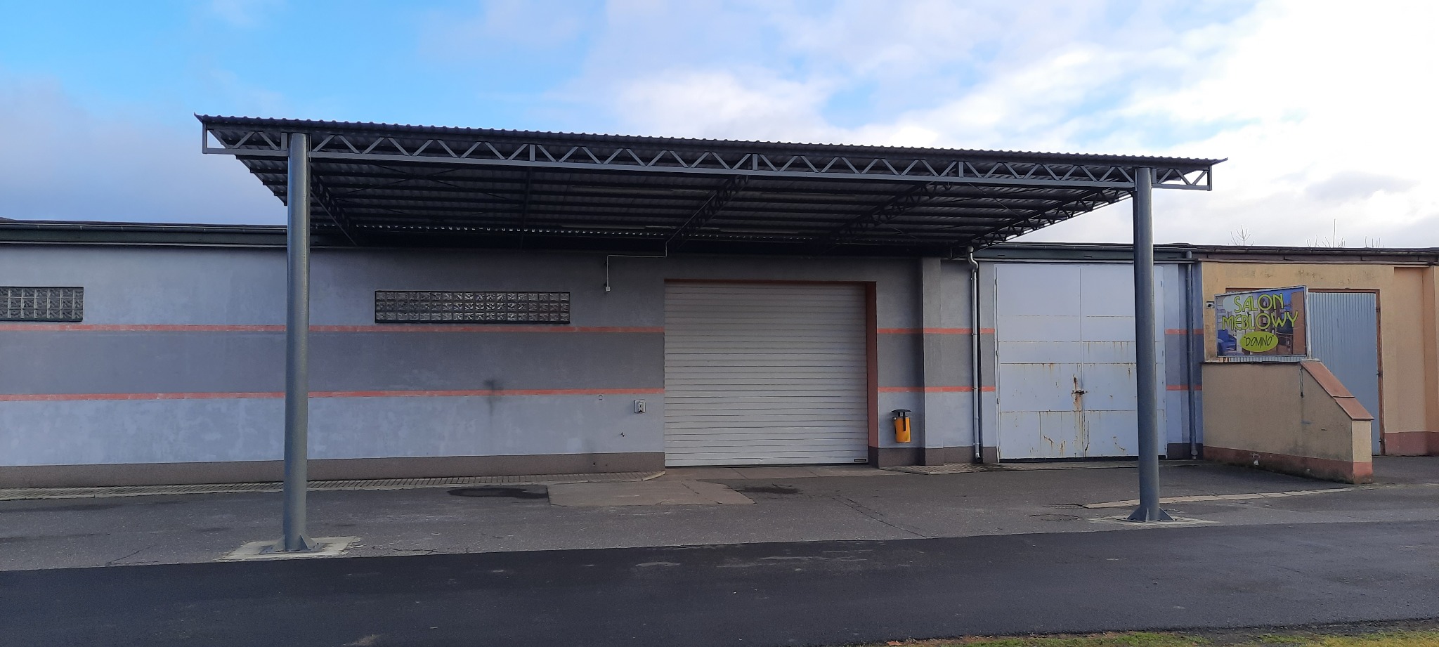 Szary budynek z bramą garażową i zadaszeniem wspartym na słupach. Na budynku widoczne okna z luksferów i reklama salonu meblowego.