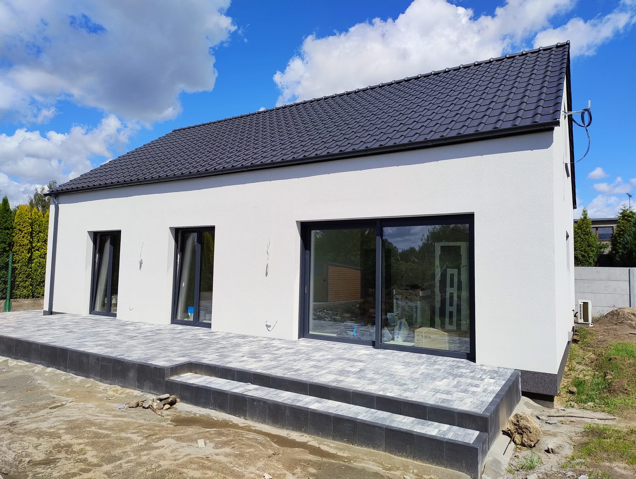 Nowoczesny, parterowy dom z szarą dachówką i wybrukowanym tarasem w Zaniemyślu. Widok z zewnątrz, w tle błękitne niebo z chmurami. Minimalistyczna architektura.
