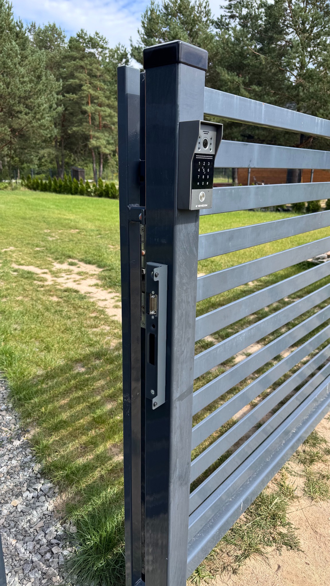 Szary wideodomofon z klawiaturą numeryczną zamontowany na metalowej bramie posesji, widok z bliska na zamek i panel. Trawa i drzewa w tle.