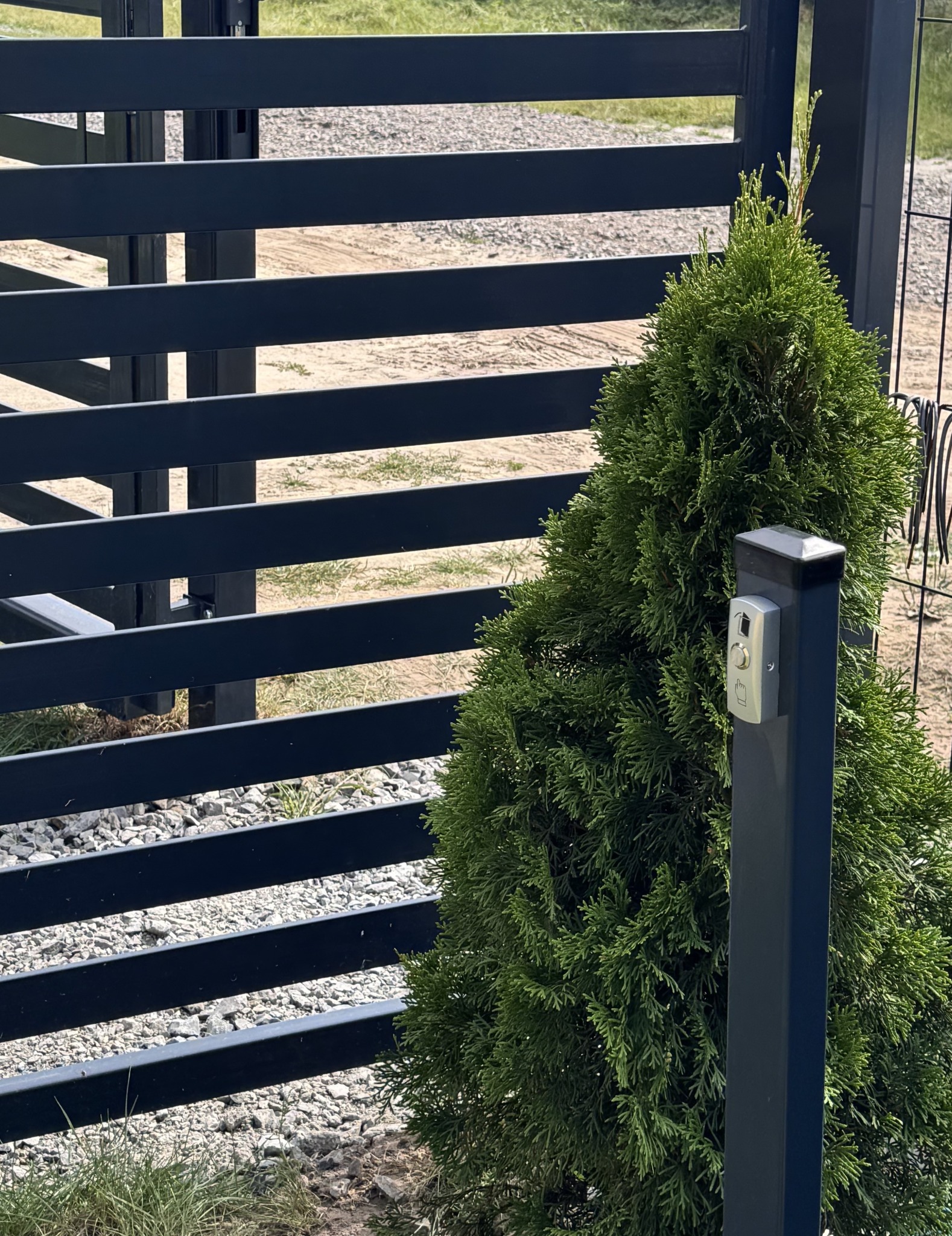 Zamontowany wideodomofon przy nowoczesnej, poziomej bramie wjazdowej, obok ozdobnego krzewu. Srebrny panel z przyciskiem dzwonka.