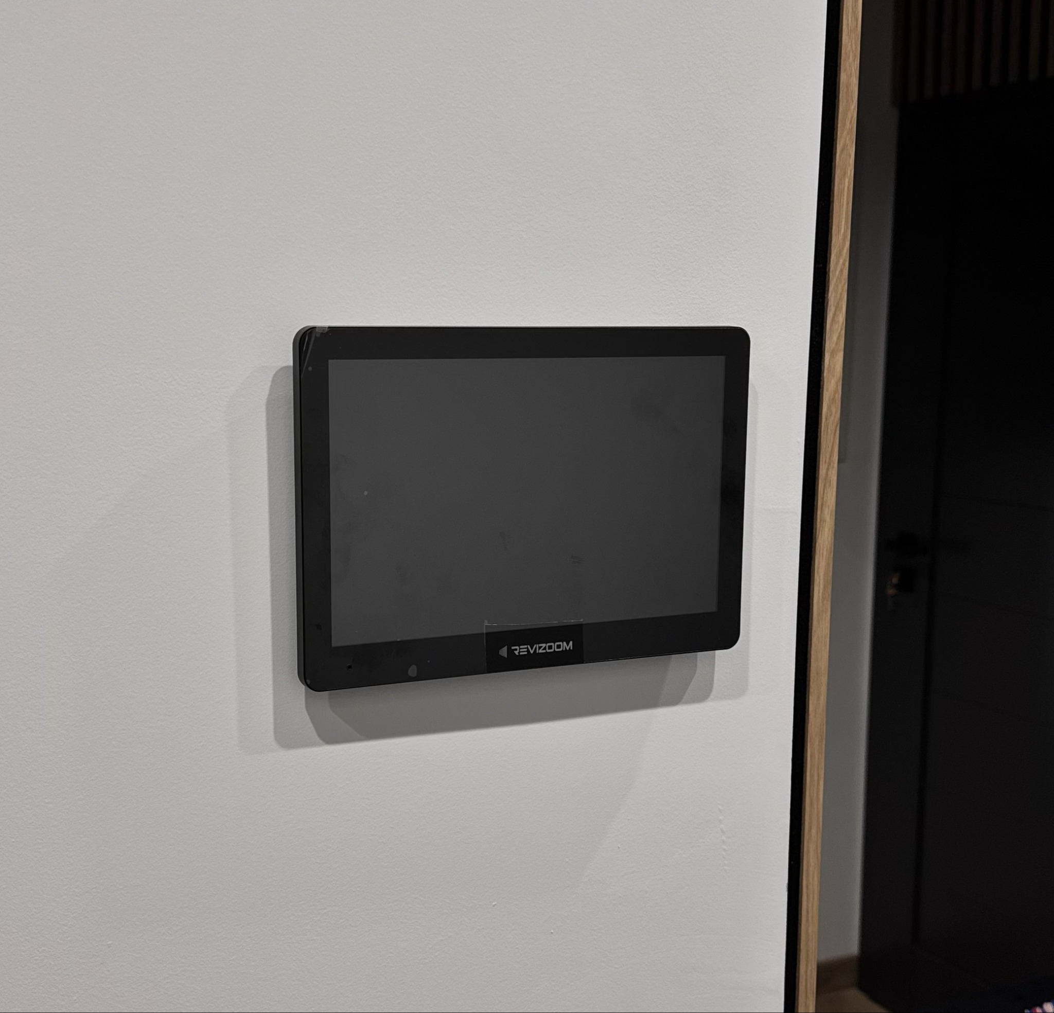 Zamontowany na ścianie, czarny panel wideodomofonu marki Revizoom, z widocznym fragmentem drzwi w tle. Minimalistyczny design.
