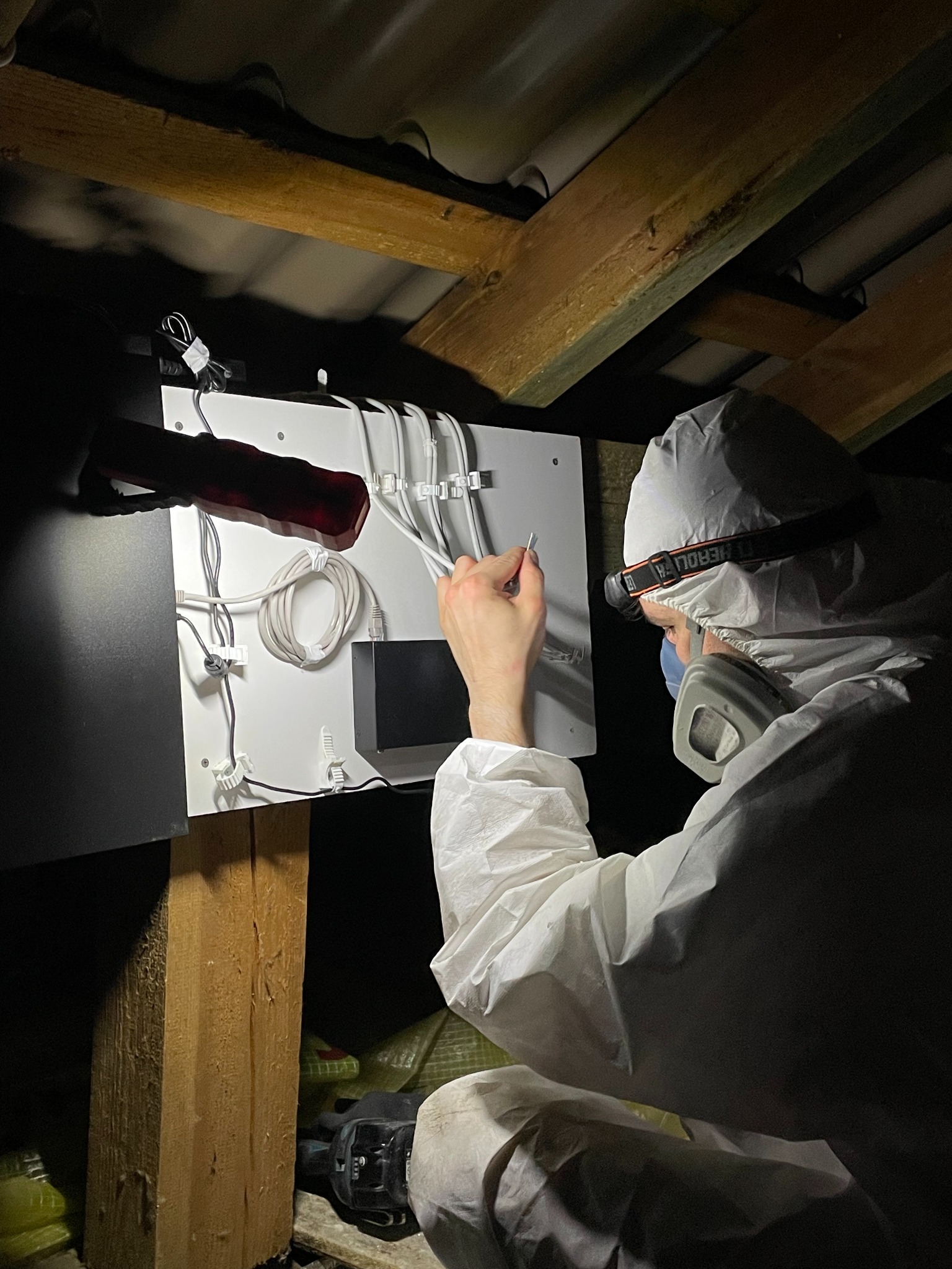 Instalator w kombinezonie ochronnym montuje okablowanie systemu monitoringu na białej płycie w przestrzeni poddasza z widocznymi elementami konstrukcji dachu.