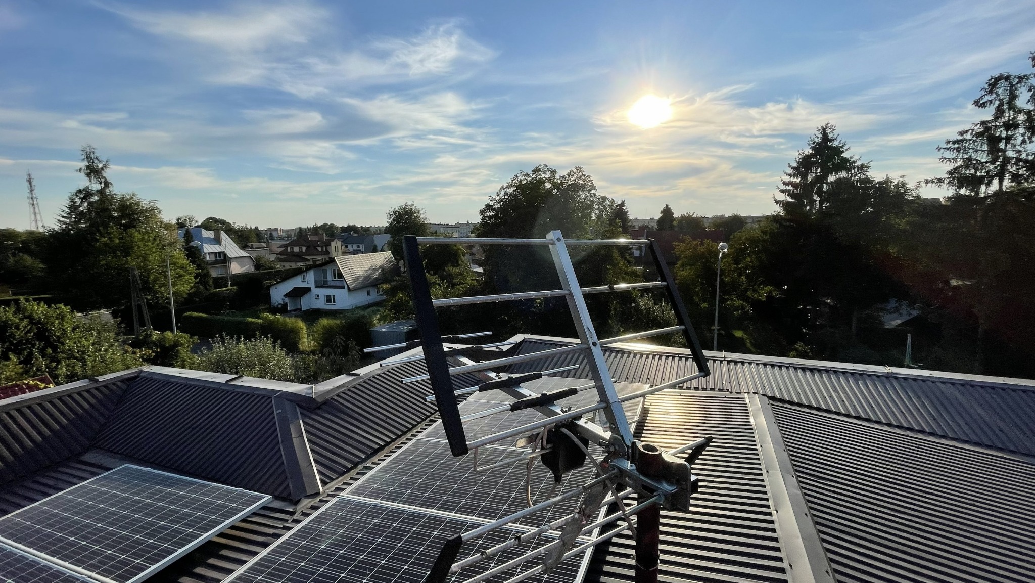 Antena telewizyjna zamontowana na dachu z blachy trapezowej, obok paneli fotowoltaicznych. W tle zabudowa jednorodzinna i błękitne niebo z promieniami słońca.