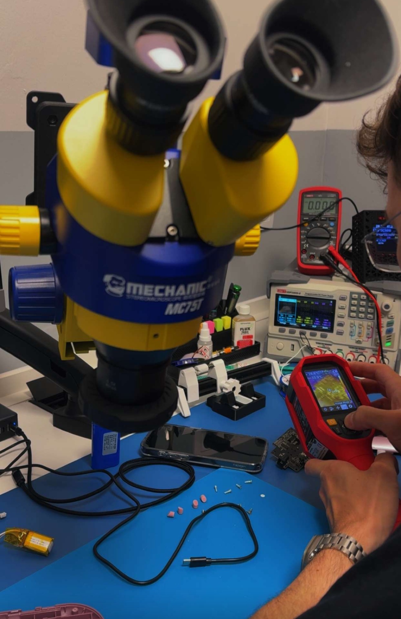 Serwis elektroniki: Mikroskop MECHANIC MC75T nad smartfonem, oscyloskop, multimetr i kamera termowizyjna w dłoniach serwisanta. Precyzyjna diagnostyka i naprawa.