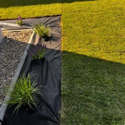 NAK SOLUTIONS SPÓŁKA Z OGRANICZONĄ ODPOWIEDZIALNOŚCIĄ - Fragment ogrodu: świeżo ułożona trawa z rolki sąsiaduje z rabatą wyłożoną czarną agrowłókniną, na której posadzone są rośliny w doniczkach. Widoczne obrzeże rabaty.