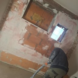 LUXO - uslugi budowlane - Prace tynkarskie wewnątrz budynku: murarz aplikuje tynk maszynowy na ścianę z cegły, widoczne okno i fragment sufitu. Praca w toku, surowy stan wykończenia.