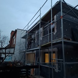 LUXO - uslugi budowlane - Prace ociepleniowe na elewacji domu jednorodzinnego z użyciem rusztowania. Widoczna warstwa izolacyjna i pracownik w trakcie montażu. Ujęcie z dołu, pochmurne niebo.