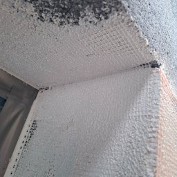 LUXO - uslugi budowlane - Narożnik ściany z siatką i warstwą kleju, przygotowany do dalszych prac wykończeniowych. Widoczne elementy styropianu i surowego betonu. Detal z bliska.