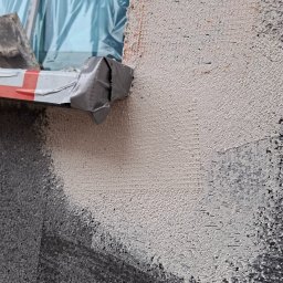 LUXO - uslugi budowlane - Fragment ocieplanej ściany z siatką i nałożonym klejem, widoczne narzędzie z taśmą. Detal prac elewacyjnych, bliski kadr na strukturę materiałów i etap ocieplania.