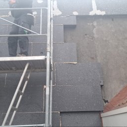 LUXO - uslugi budowlane - Prace ociepleniowe elewacji budynku z użyciem czarnych płyt styropianowych, widoczne rusztowanie i fragment dachu z czerwonej dachówki. Etap montażu izolacji.