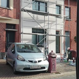 LUXO - uslugi budowlane - Dom w trakcie ocieplania elewacji, widoczne rusztowania i pracownik przy oknie. Obok stoi srebrny samochód i czerwona skrzynka pocztowa. Częściowo ocieplona ściana.