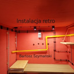 A1ELEKTRO - Retro instalacja elektryczna z czarnymi przewodami i żółtymi rurami na czerwonym tle. Widoczne oświetlenie punktowe i drewniane półki.