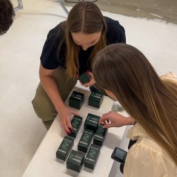 PIONA Studio Oskar Makarski - Dwie osoby układają pudełka z produktami marki Shinow na białym stole w studio fotograficznym. Widoczny sprzęt oświetleniowy i butelka z płynem.