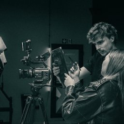 PIONA Studio Oskar Makarski - Czarno-białe zdjęcie z planu filmowego: kamera Sony na statywie, reżyser z aktorką omawiają scenariusz. W tle ciemne pomieszczenie i znak ewakuacyjny.