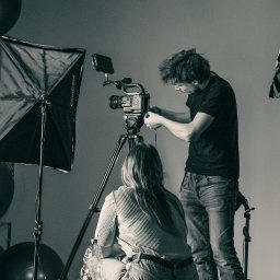 PIONA Studio - Czarno-białe zdjęcie zza kulis: mężczyzna reguluje kamerę na statywie, obok softbox CineGEN, tablica z napisem SMART, kobieta siedzi na podłodze. Studio.