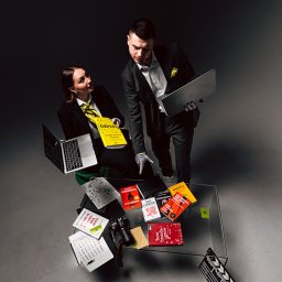 PIONA Studio - Kreatywny zespół z laptopami i książkami o biznesie na szklanym stole. Nietypowe ujęcie z góry, ciemne tło, studyjne oświetlenie. Koncepcja edukacji i rozwoju w biznesie.