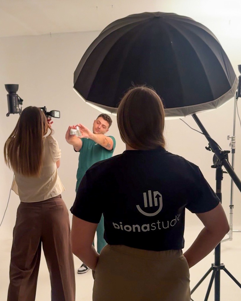 Sesja zdjęciowa w studio: mężczyzna w zielonym stroju prezentuje produkt, fotografka robi zdjęcia, a osoba z logo 'bionastudio' obserwuje.