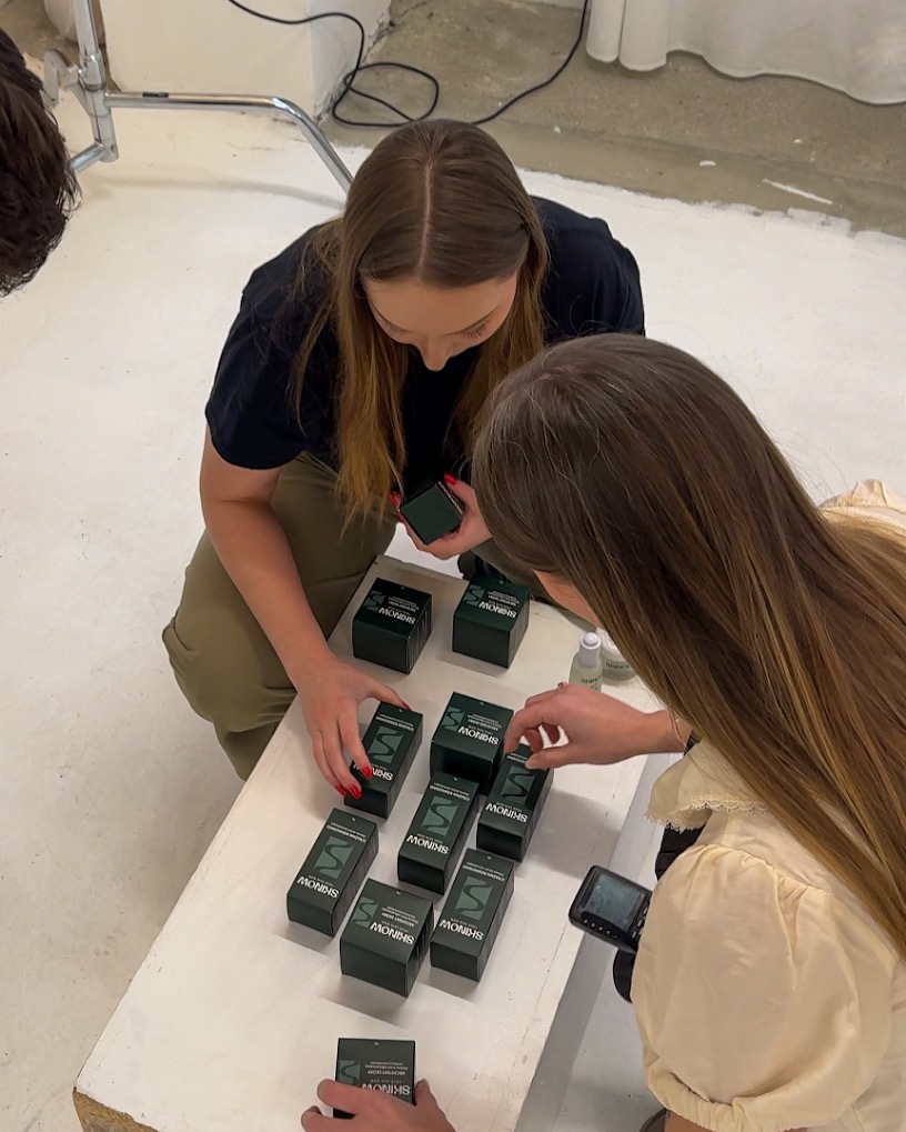 Dwie osoby układają pudełka z produktami marki Shinow na białym stole w studio fotograficznym. Widoczny sprzęt oświetleniowy i butelka z płynem.