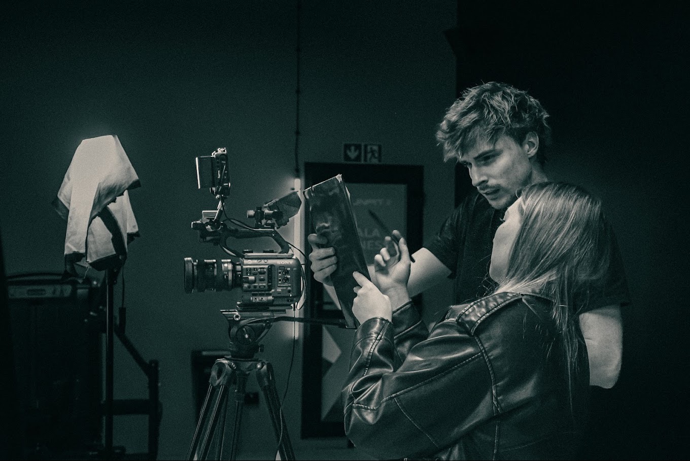 Czarno-białe zdjęcie z planu filmowego: kamera Sony na statywie, reżyser z aktorką omawiają scenariusz. W tle ciemne pomieszczenie i znak ewakuacyjny.