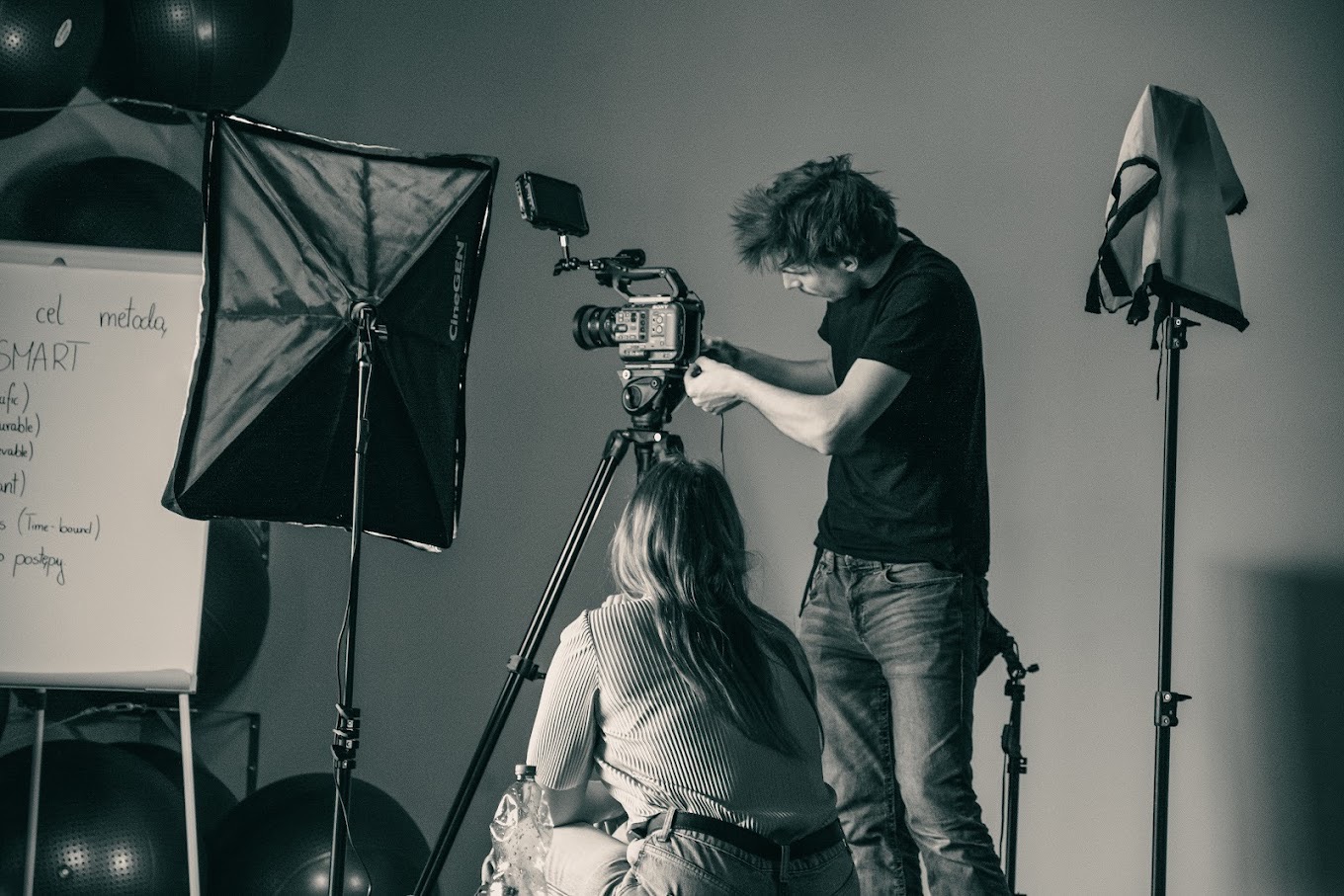 Czarno-białe zdjęcie zza kulis: mężczyzna reguluje kamerę na statywie, obok softbox CineGEN, tablica z napisem SMART, kobieta siedzi na podłodze. Studio.