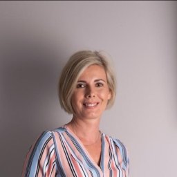 Sylwia Zaleska - Portret uśmiechniętej kobiety z krótkimi blond włosami, ubrana w bluzkę w pastelowe paski, na jasnoszarym tle. Zdjęcie biznesowe, budzące zaufanie.