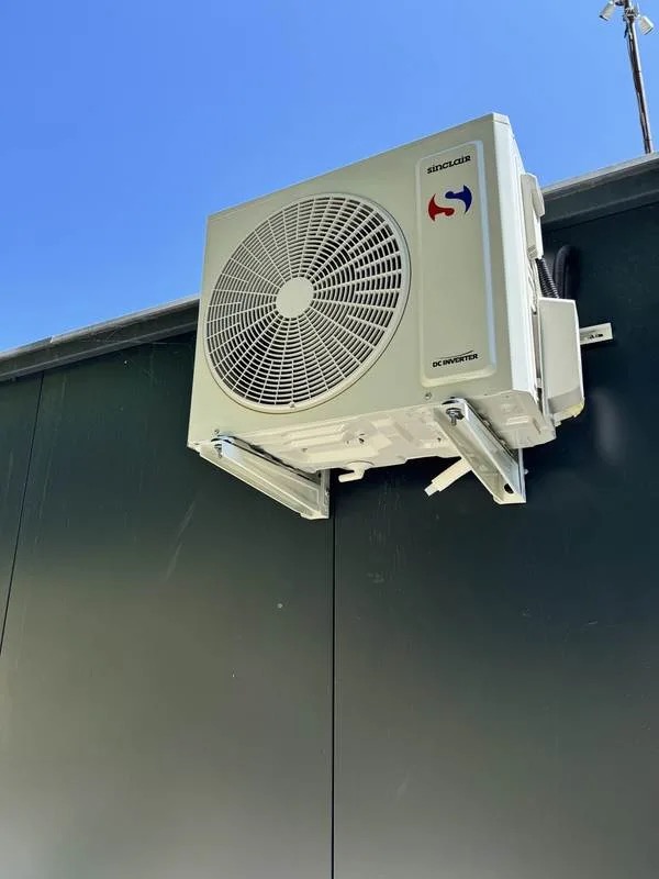 Biała jednostka zewnętrzna klimatyzacji Sinclair zamontowana na ciemnej ścianie budynku, widoczna na tle błękitnego nieba. Metalowe wsporniki mocujące.