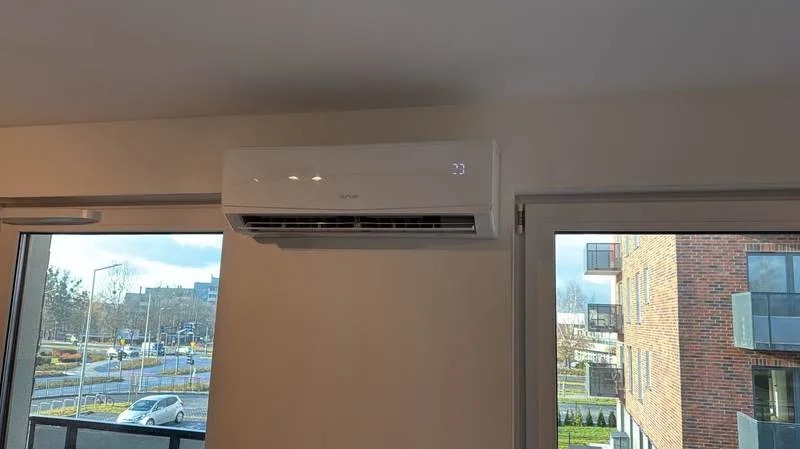 Biały klimatyzator ścienny z wyświetlaczem temperatury (23°C) zamontowany w jasnym wnętrzu, widok z okna na budynek mieszkalny.