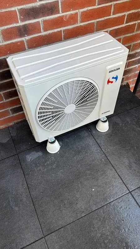 Biała jednostka zewnętrzna klimatyzacji Sinclair na szarych płytkach i ceglanej ścianie. Urządzenie stoi na czterech białych, stożkowych nóżkach.