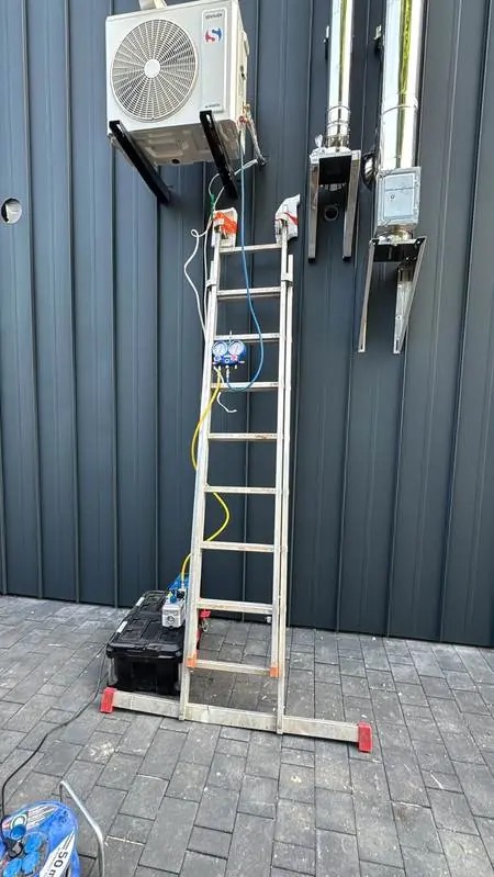 Montaż klimatyzacji zewnętrznej na szarej elewacji. Aluminiowa drabina z narzędziami serwisowymi. Widoczne rury wentylacyjne. Kostka brukowa na podłożu.