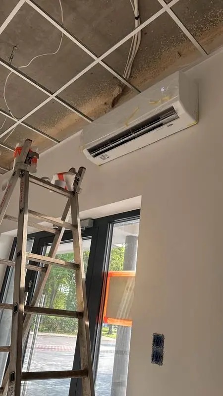Montaż klimatyzatora na ścianie, widoczna drabina i sufit z instalacjami. Urządzenie jeszcze w folii ochronnej, w tle okno i fragment ulicy.