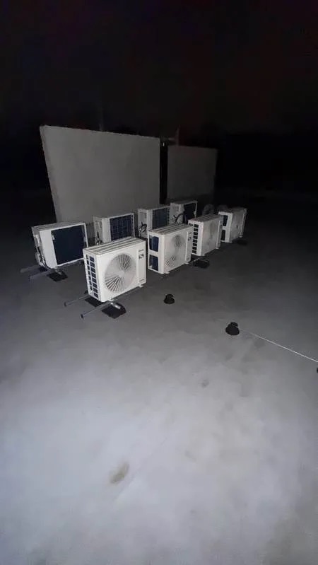 Rząd białych jednostek zewnętrznych klimatyzacji na dachu, widok z góry w słabym oświetleniu, z widocznymi wspornikami montażowymi i elementami instalacji.